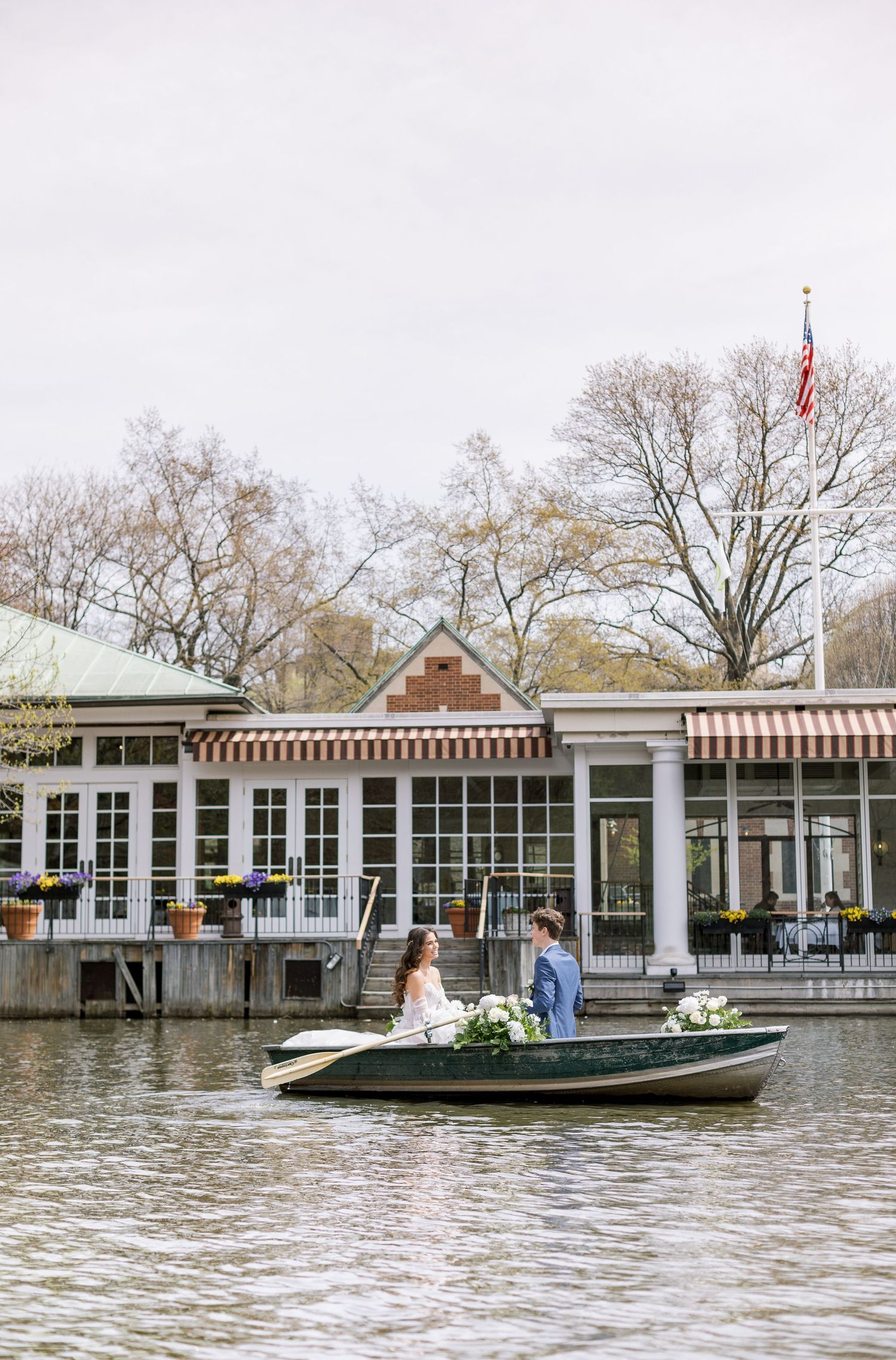 Wedding couple in rowboat near lakeside restaurant with striped awnings.