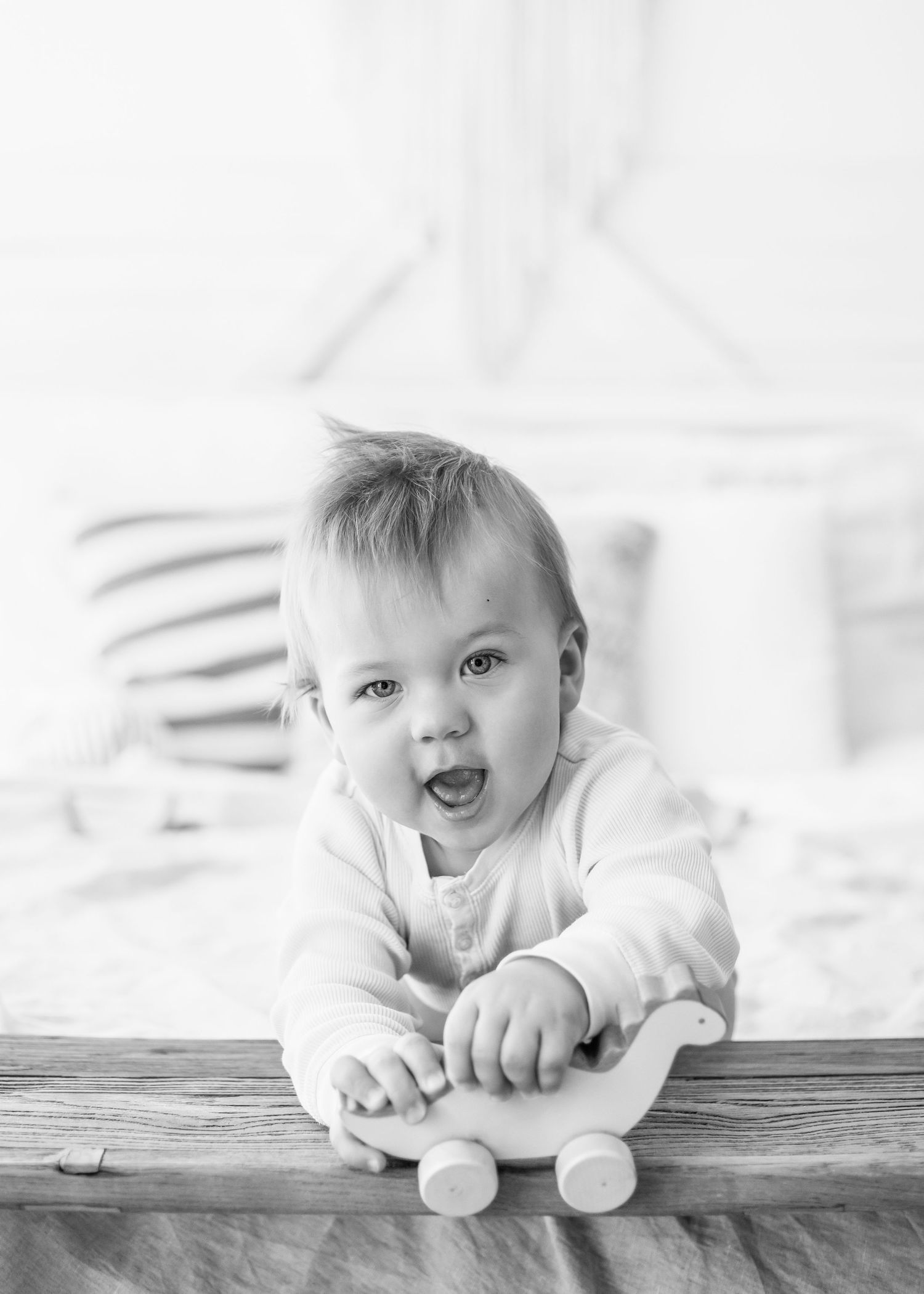 A happy baby in white plays with a wooden toy while lying on their stomach.