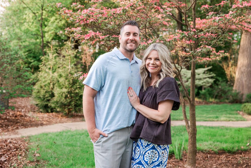 Couple stands together in garden setting with pink flowering trees in background.