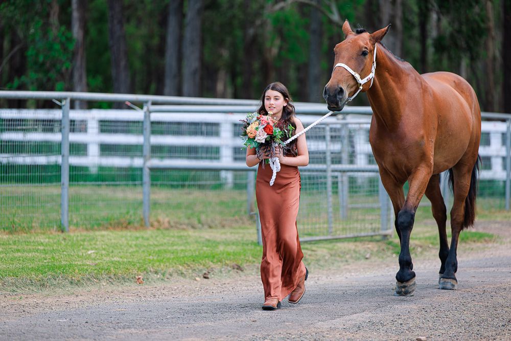 Wedding party members walk along a dirt path with a brown horse at a rustic venue.