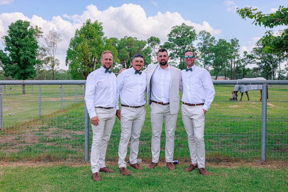 Wedding party in all-white attire poses together on grassy field near white fence.