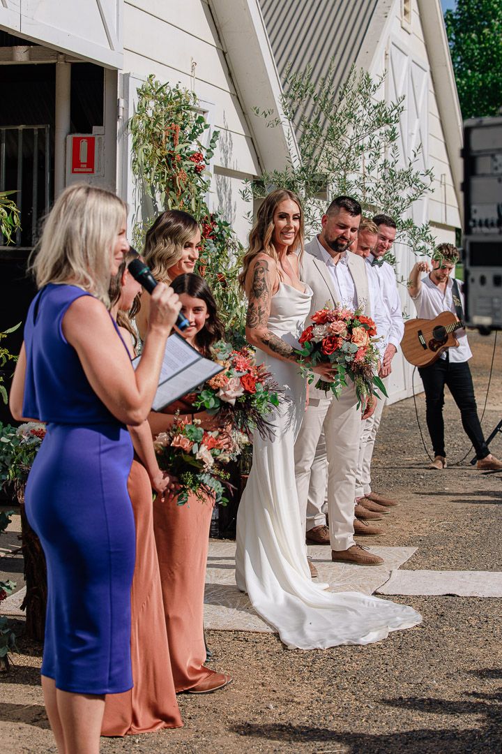 Wedding ceremony setup outside white barn with floral decorations and guests.