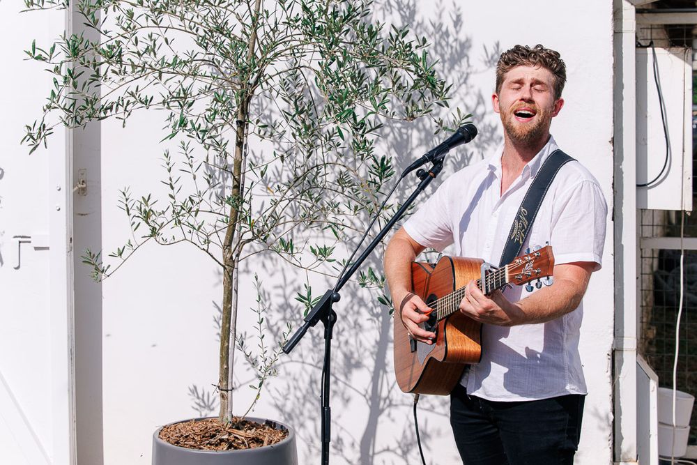 Musicians performing with acoustic guitar against white backdrop at outdoor wedding.