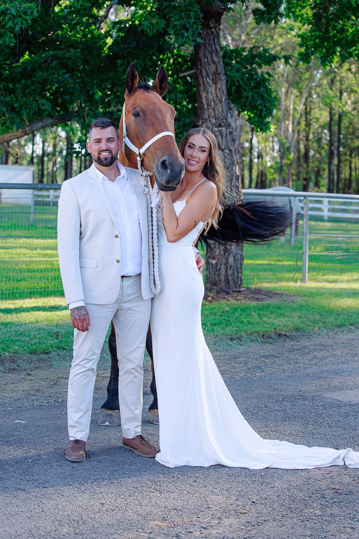 A couple poses with their brown horse on a tree-lined path during a wedding photoshoot.