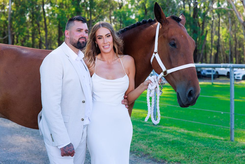 Couple in white attire poses with brown horse at outdoor wedding venue.