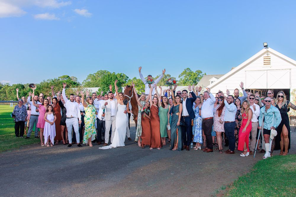 Large wedding party group photo in front of a white chapel on a sunny day.