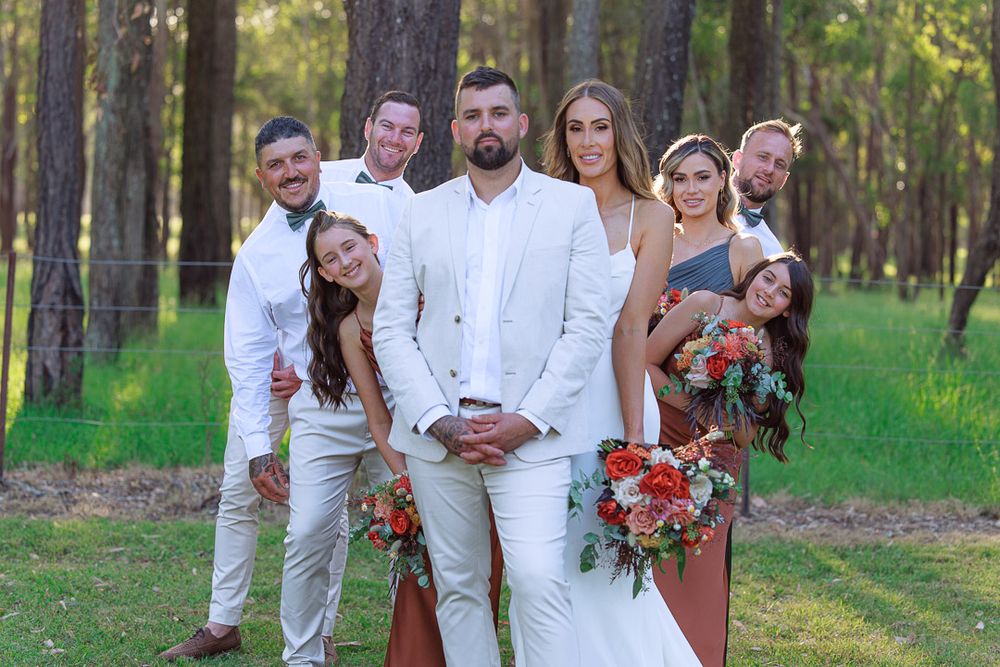 Wedding party posing together in white and burgundy attire among pine trees.