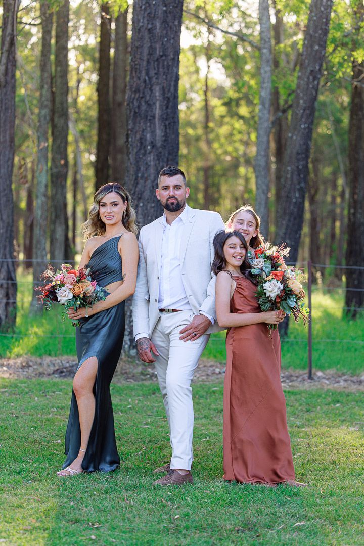 Three people in formal attire pose with floral bouquets in a pine forest setting.