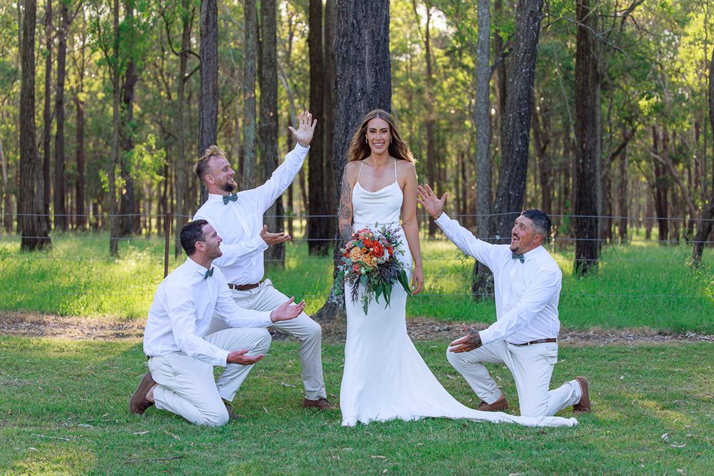 Wedding party members strike playful poses around bride in a forest setting.