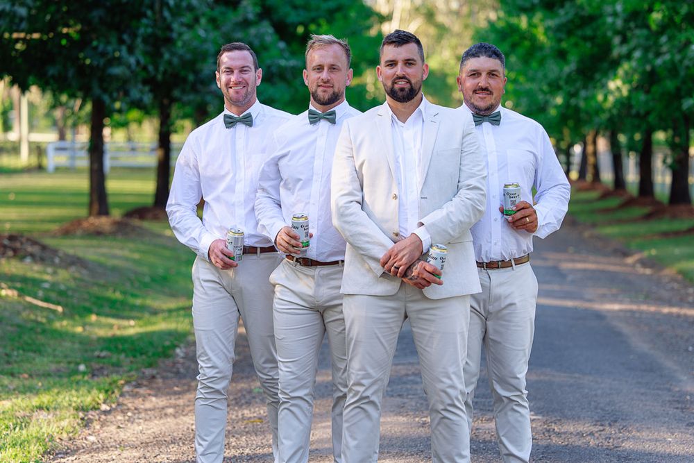 Groomsmen in light gray suits pose together on a tree-lined path during an outdoor wedding.