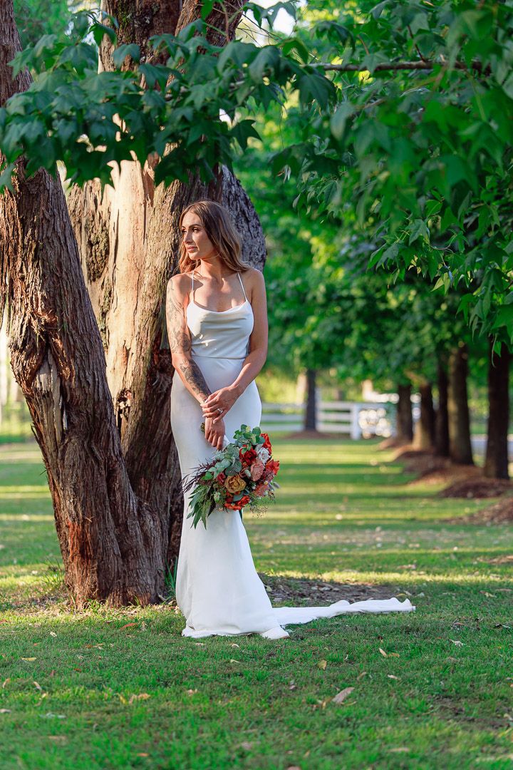 Bride in elegant white dress poses with bouquet by tree in garden setting.