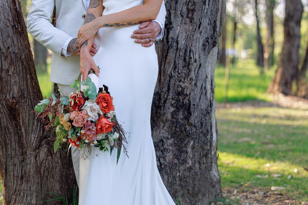 Wedding couple poses against large tree trunk with bride holding red and white bouquet.