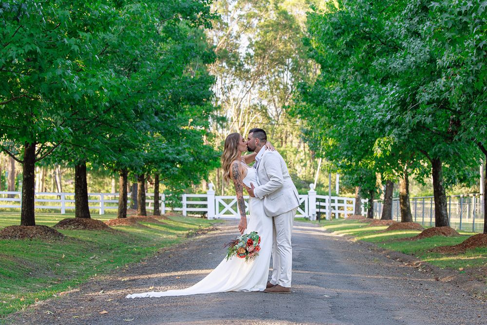 Wedding couple poses along tree-lined path with white fencing in background.