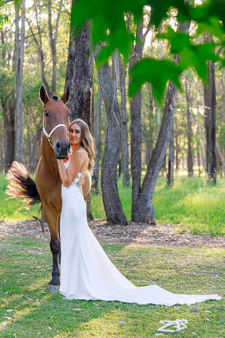 Bride in flowing white dress poses with brown horse in wooded setting.