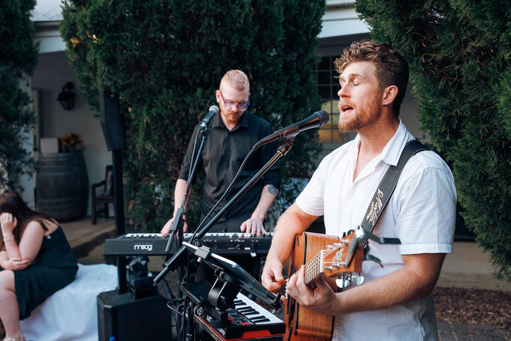 Musicians perform with guitars at outdoor venue under trees.