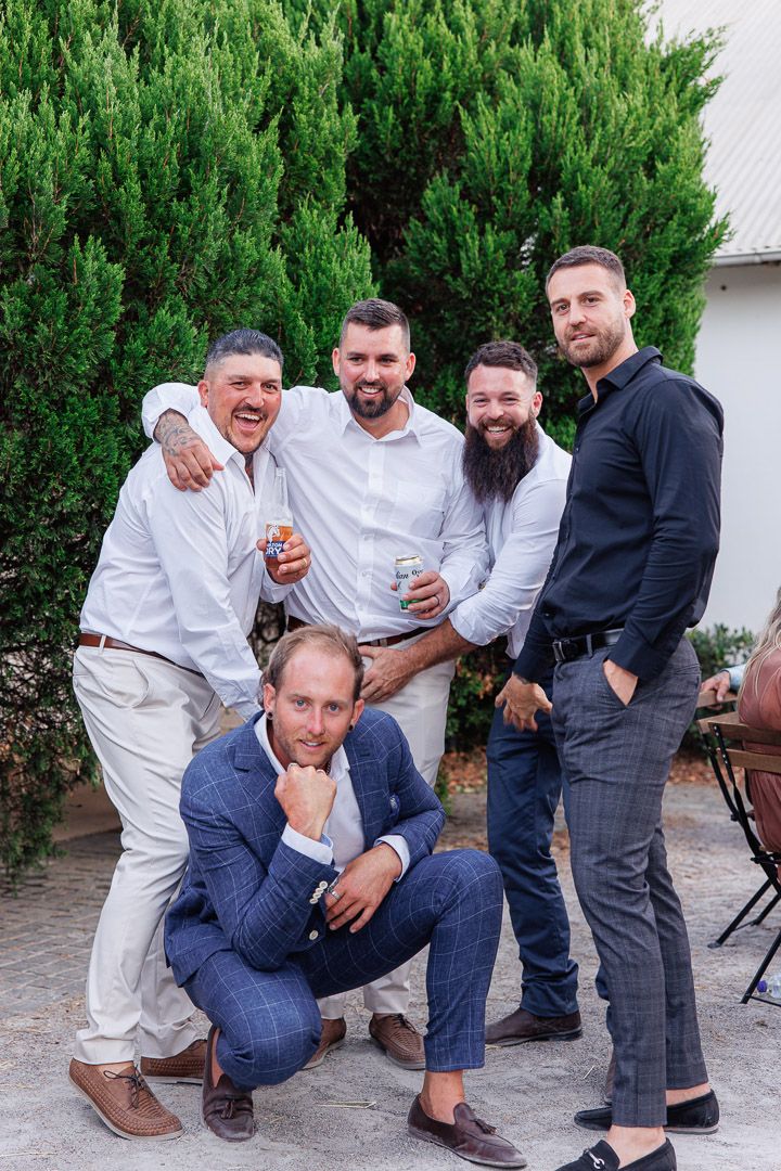 Groomsmen pose playfully together against green hedge backdrop.