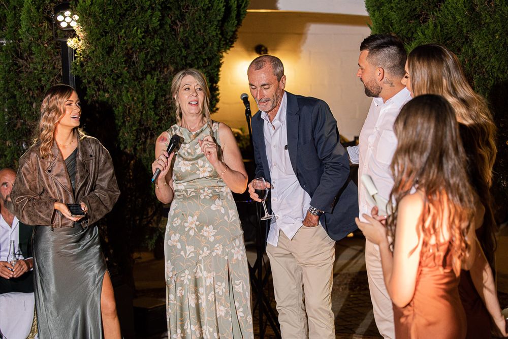 Party guests dance and celebrate at an outdoor evening reception with greenery in the background.