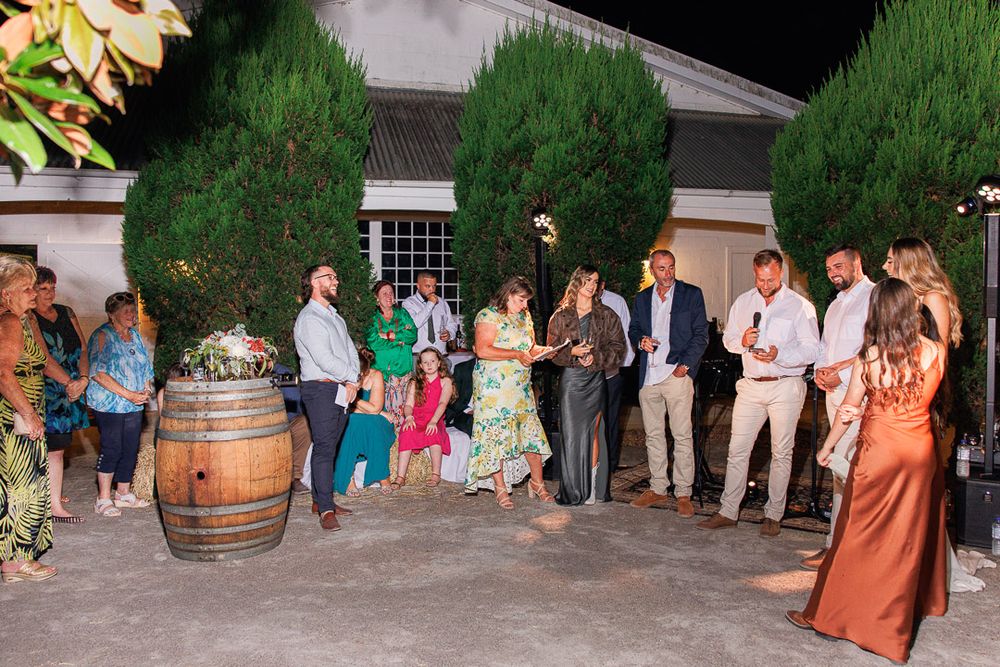 Guests gather at an outdoor party venue with wooden barrels and garden landscaping.