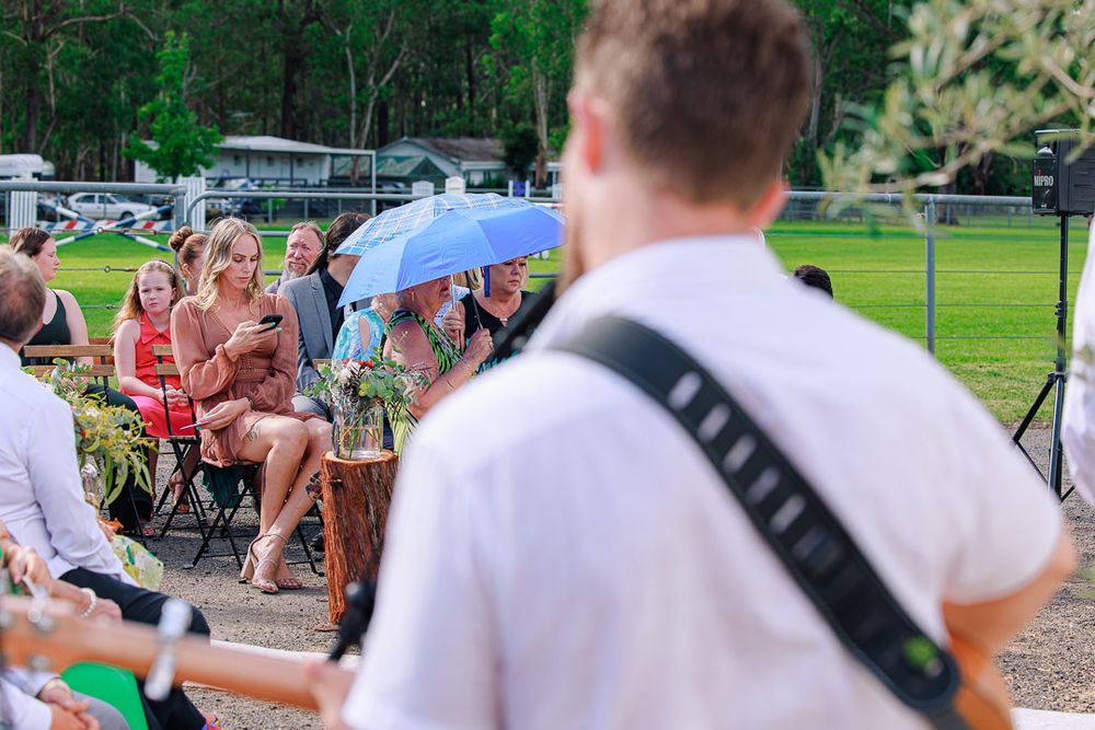 Guests seated on white chairs at an outdoor wedding ceremony with trees in the background.