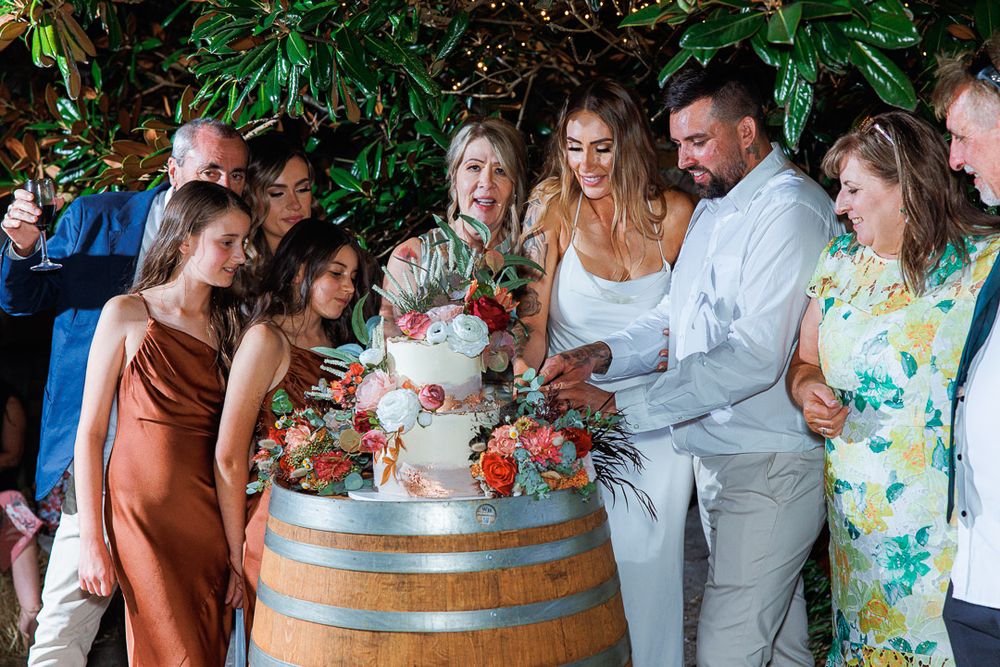 Wedding party members laughing together around a decorative barrel at outdoor reception.