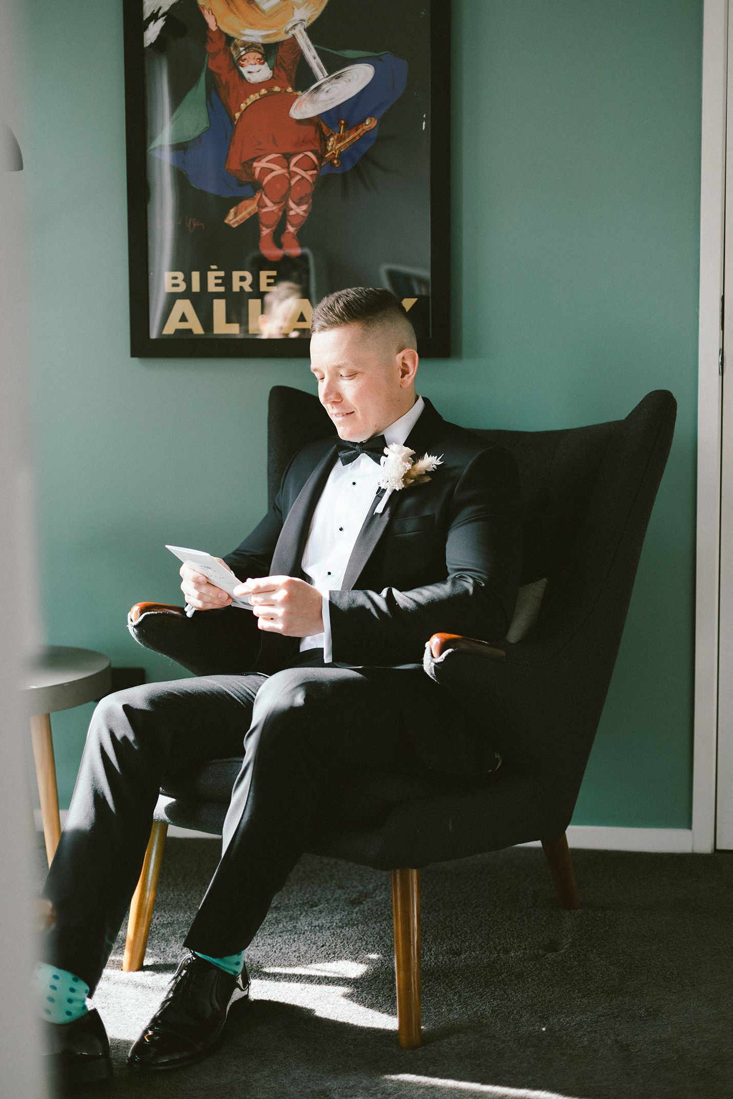A person in a tuxedo reads while seated in a wingback chair beneath vintage beer artwork.