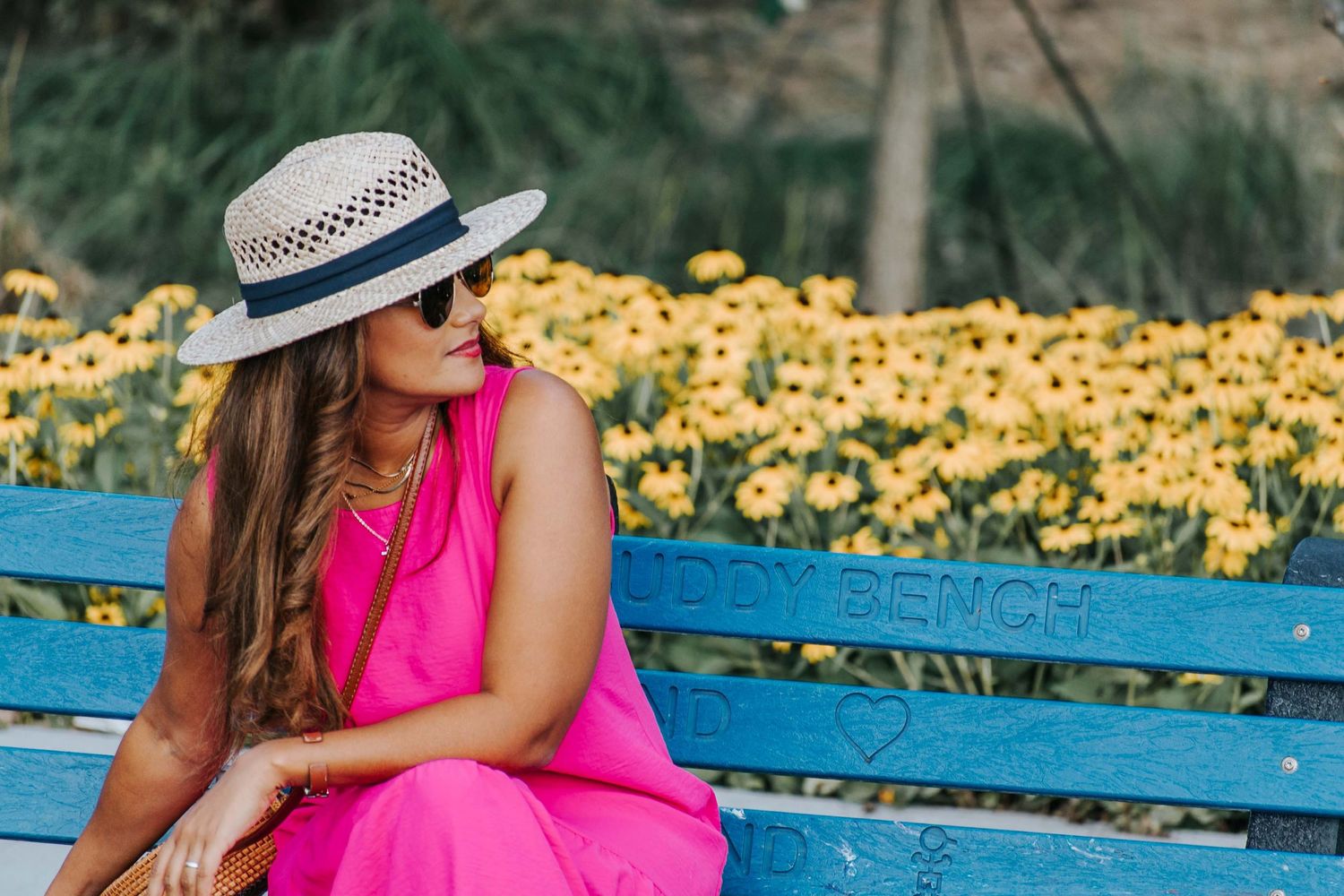 Woman in pink dress and sunhat sits on blue bench surrounded by yellow flowers.