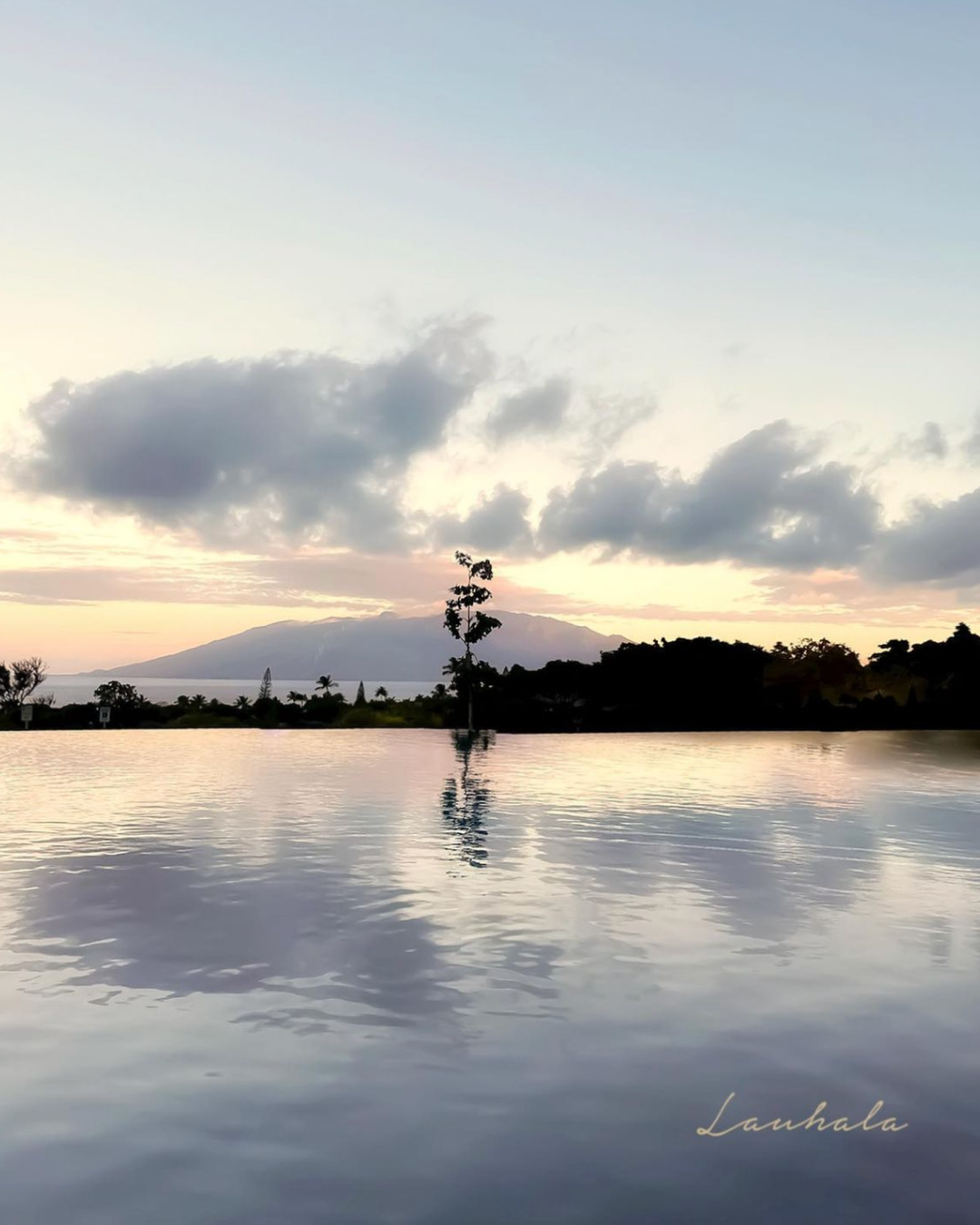Tranquil lake reflects dramatic sunset clouds with mountain silhouette in Hawaii.