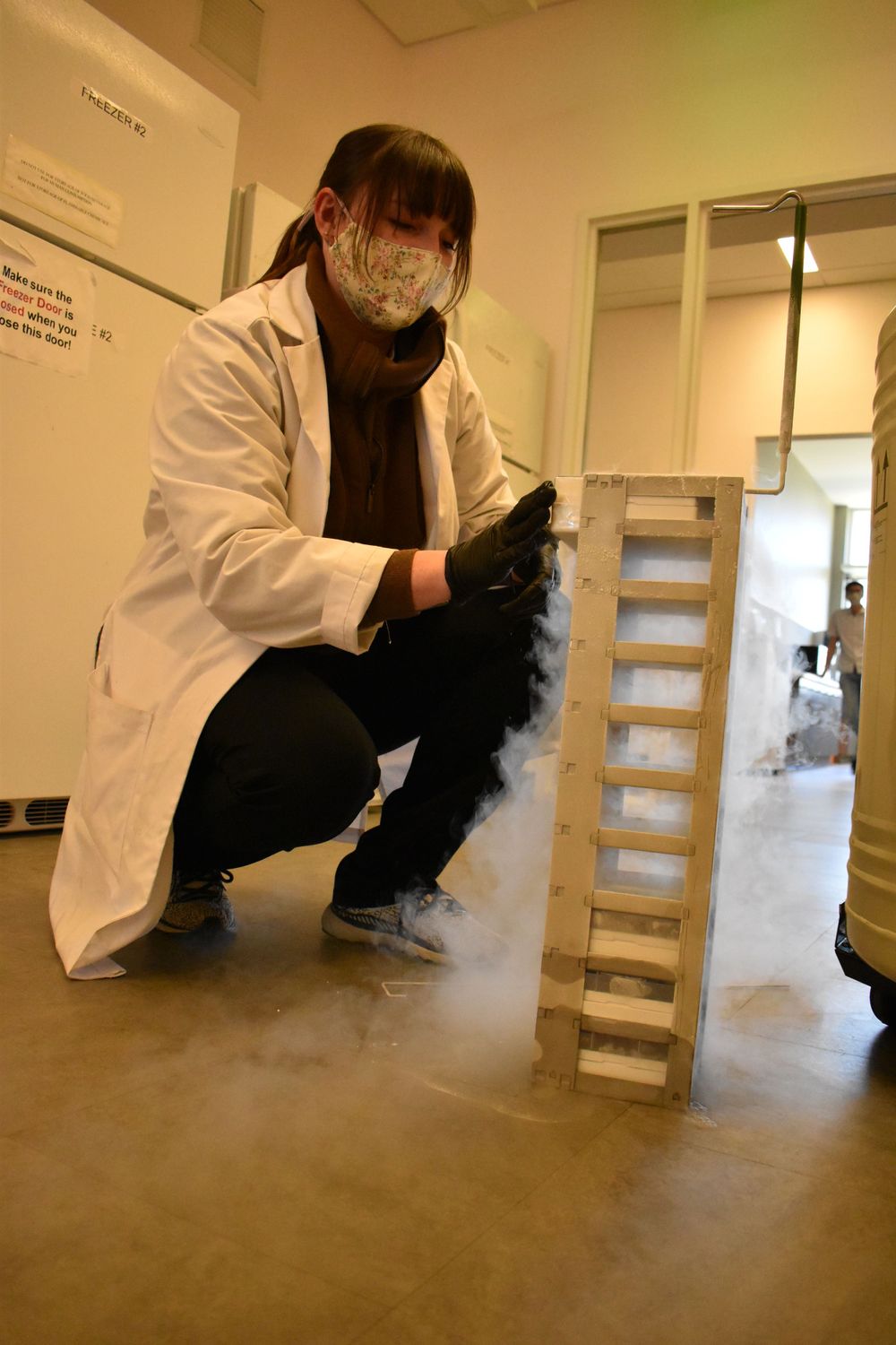 Researcher in white coat and safety gear crouches near dry ice experiment creating fog effect.