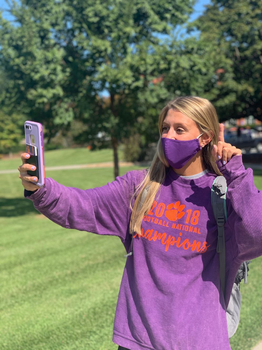 Student in purple sweatshirt takes selfie outdoors on college campus.