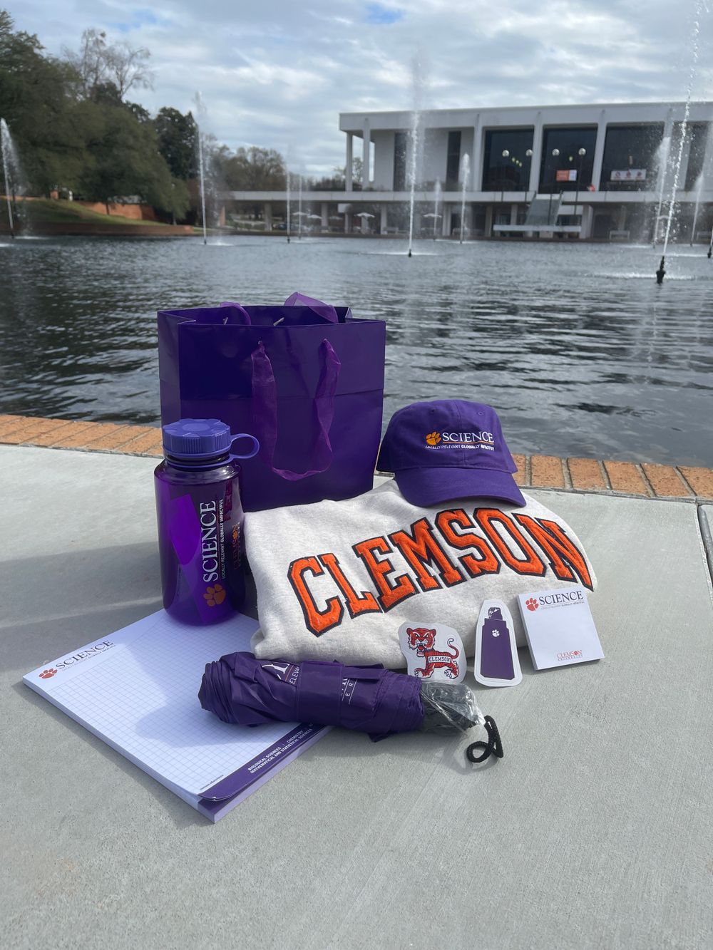 Clemson University merchandise including purple cap, water bottle and white t-shirt displayed near reflective pond.