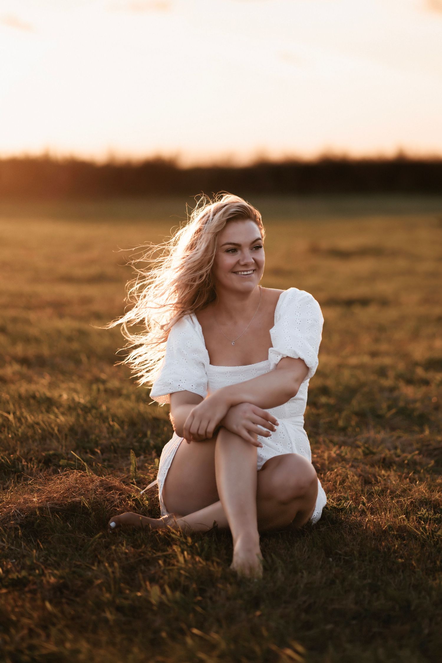 Person in white dress sits in golden field during sunset with windswept hair.