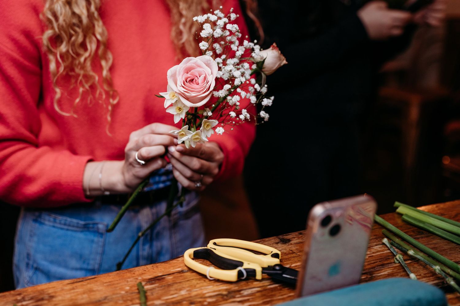 Hands arranging pink roses and baby's breath flowers on wooden table with phone visible.