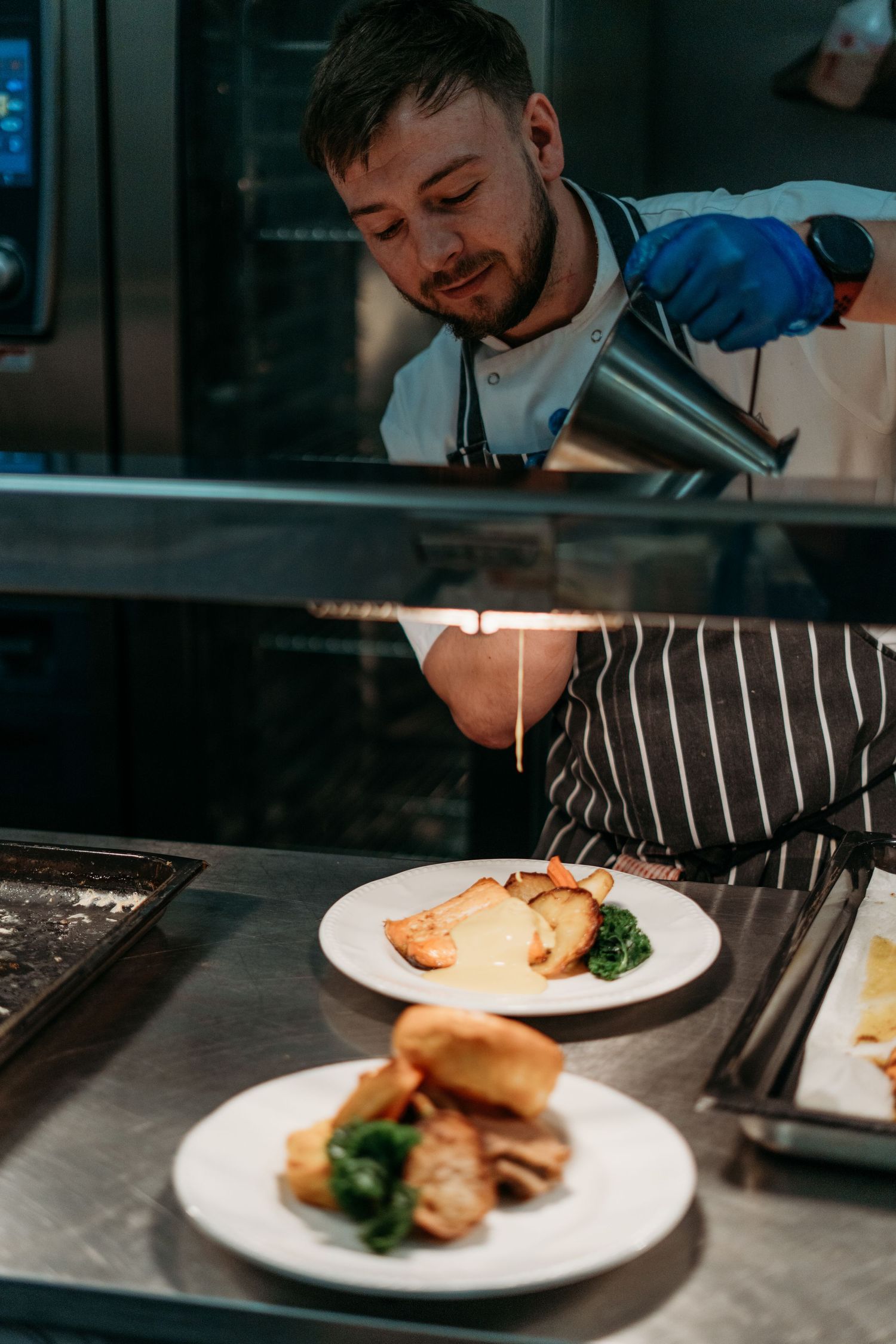 Chef in striped apron plating and garnishing elegant dinner dishes in restaurant kitchen.