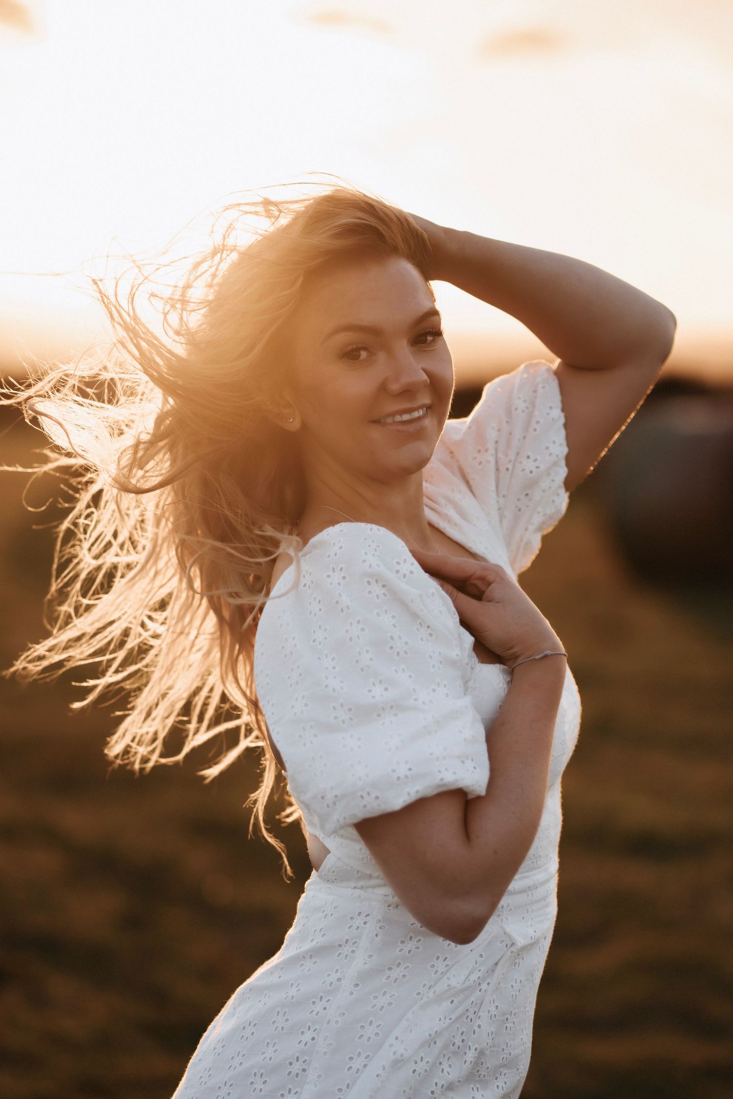Woman in white dress poses against golden sunset light with flowing hair in countryside setting.