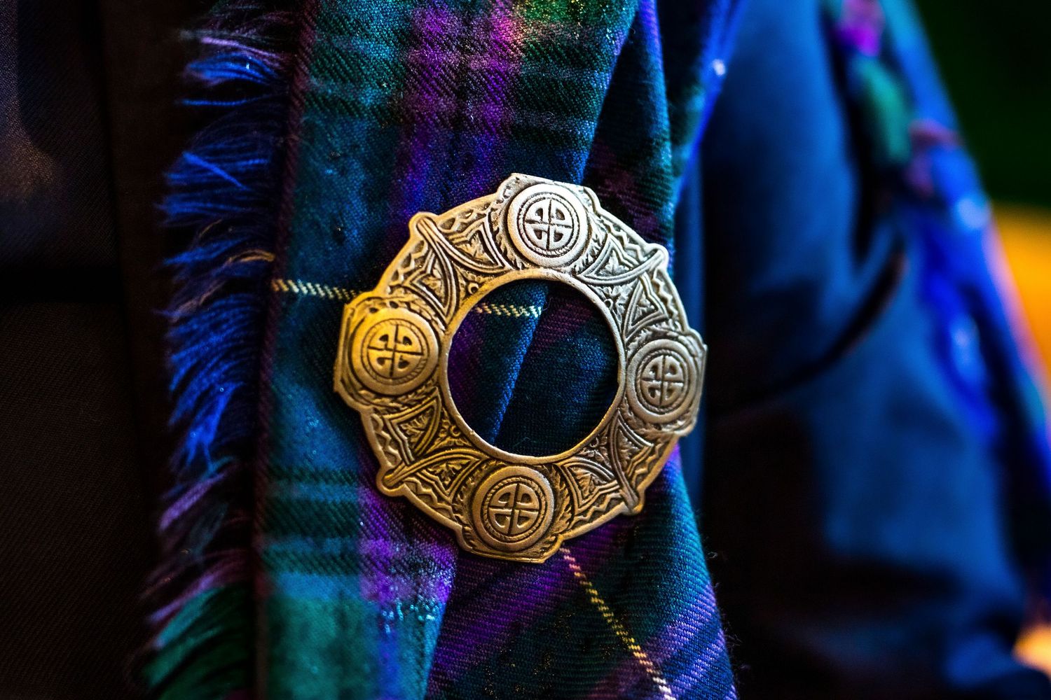 Close-up detail of an ornate Celtic-style silver brooch pin against a blue tartan fabric background.