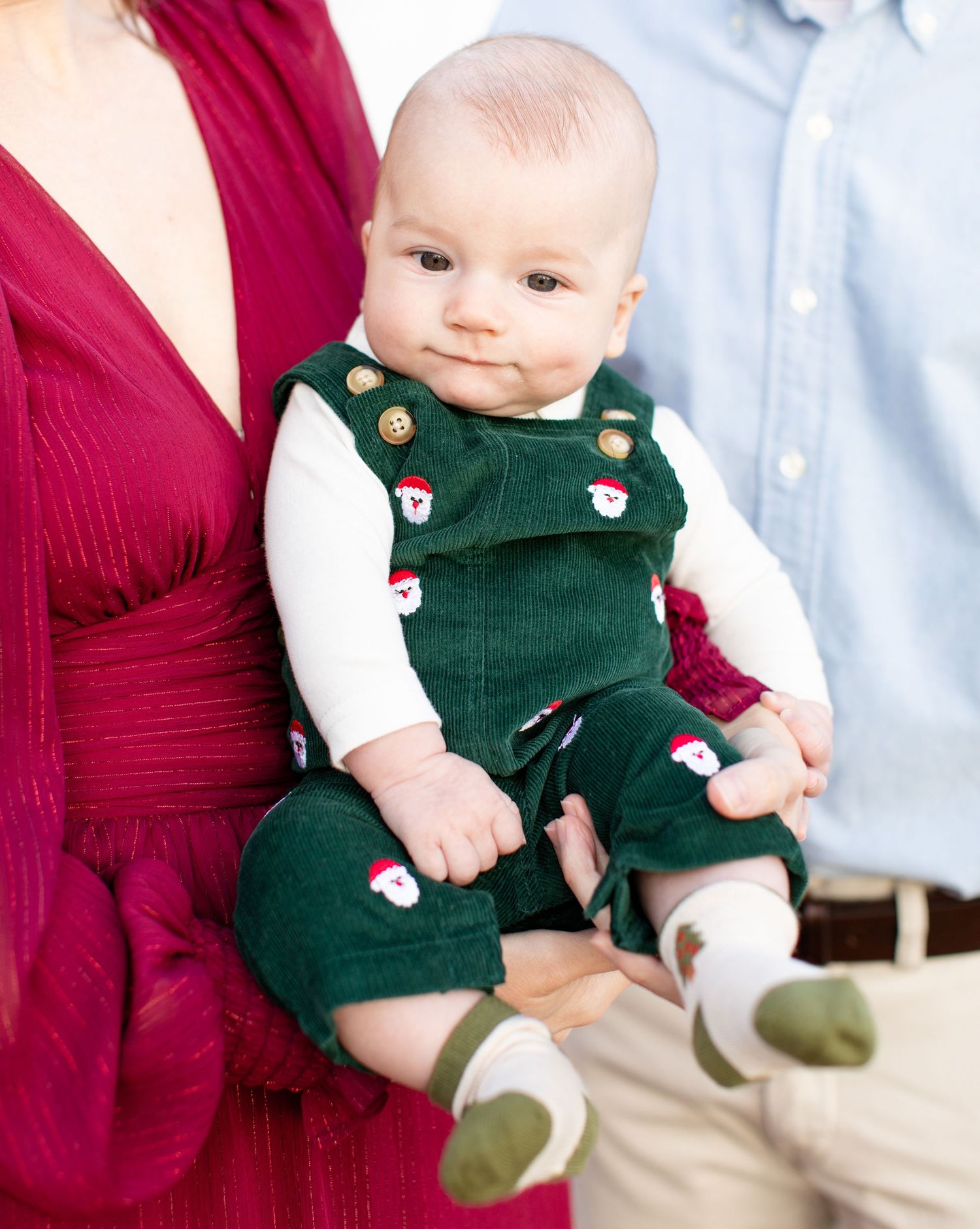 Baby wearing festive green overalls with Santa pattern between parents.