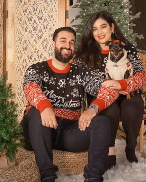 Couple in matching Christmas sweaters sits with their dog in front of a decorated tree and lace curtain.
