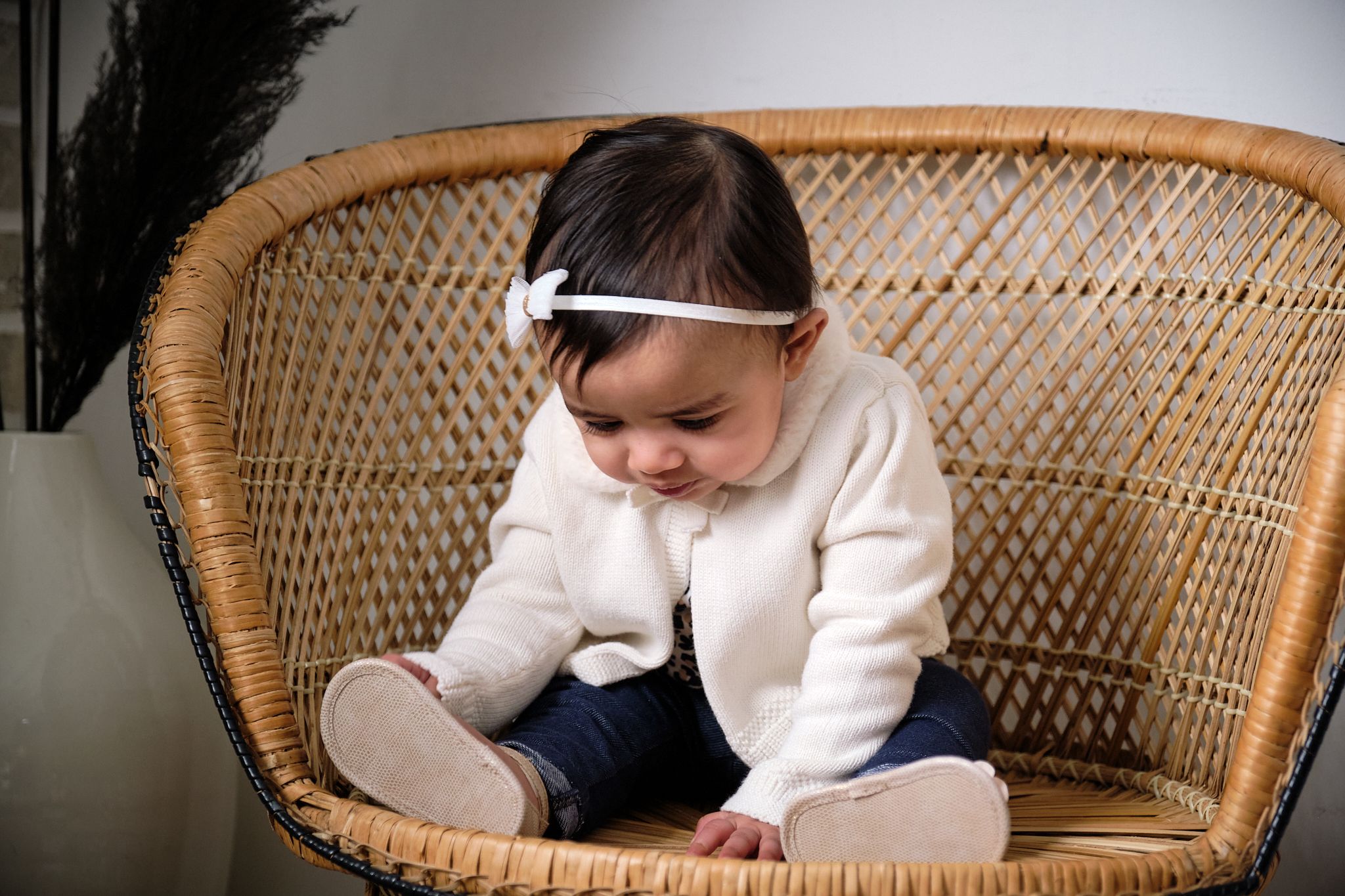 Small child wearing white sweater sits in woven rattan peacock chair looking downward.