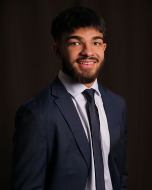 Professional headshot of person in navy suit and tie against dark background.