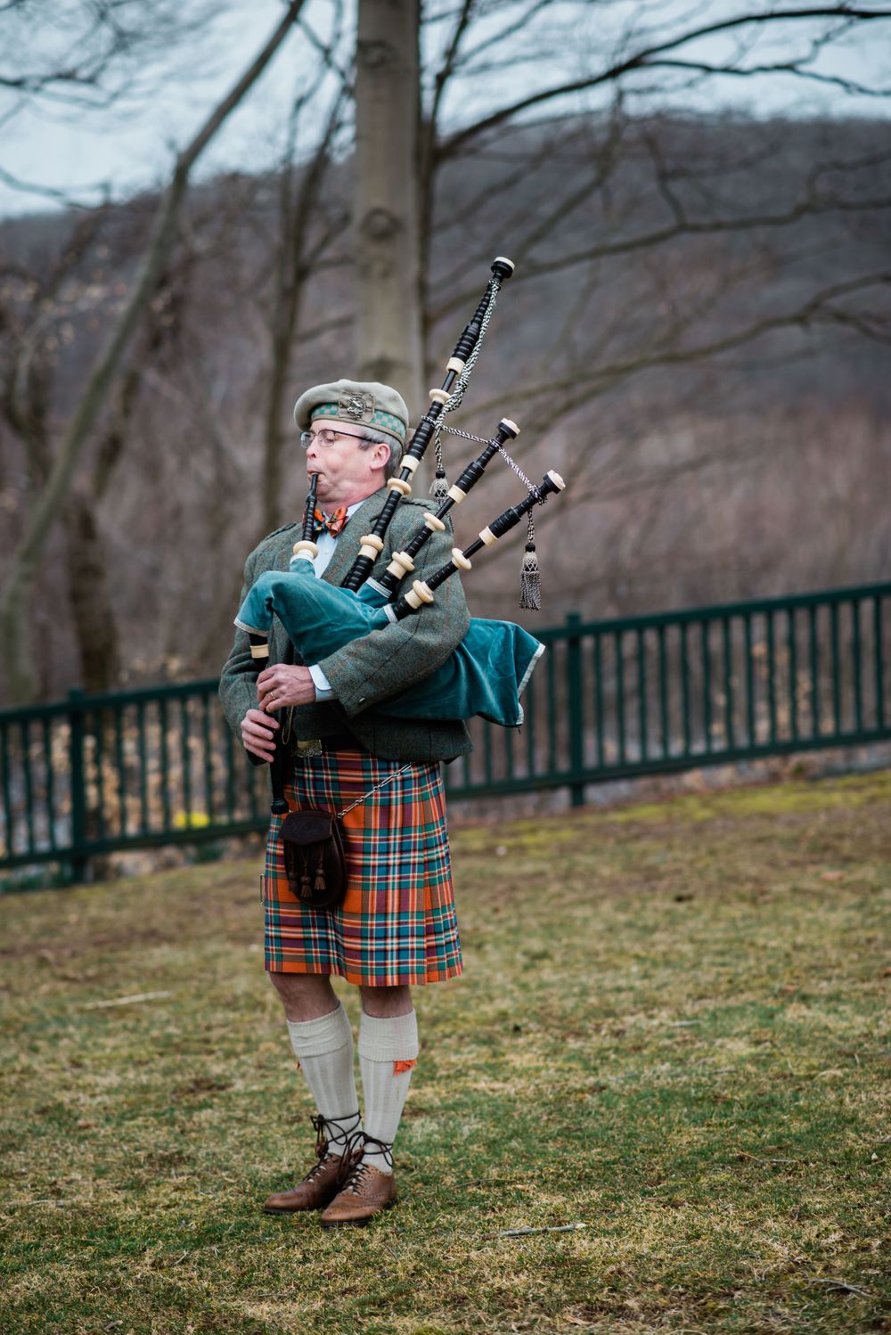 A bagpiper in traditional Scottish attire performs outdoors against a mountainous winter backdrop.
