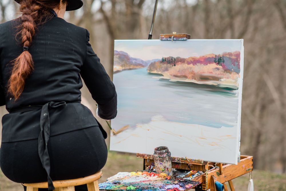 An artist paints a watercolor landscape scene while sitting outdoors.