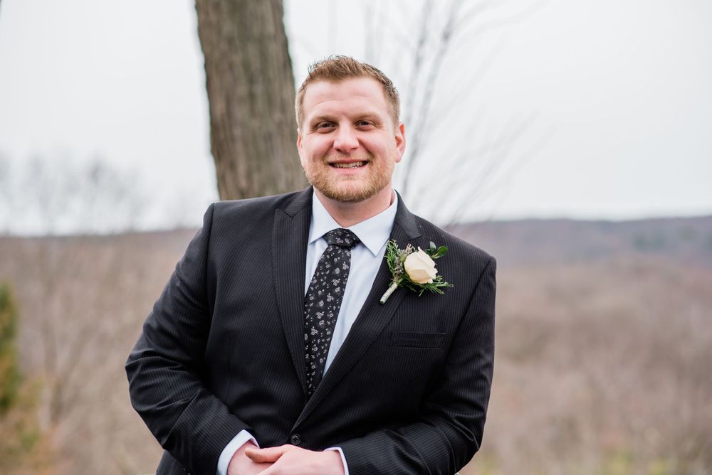 A wedding attendee in a black suit with polka dot tie stands outdoors against a mountain vista.