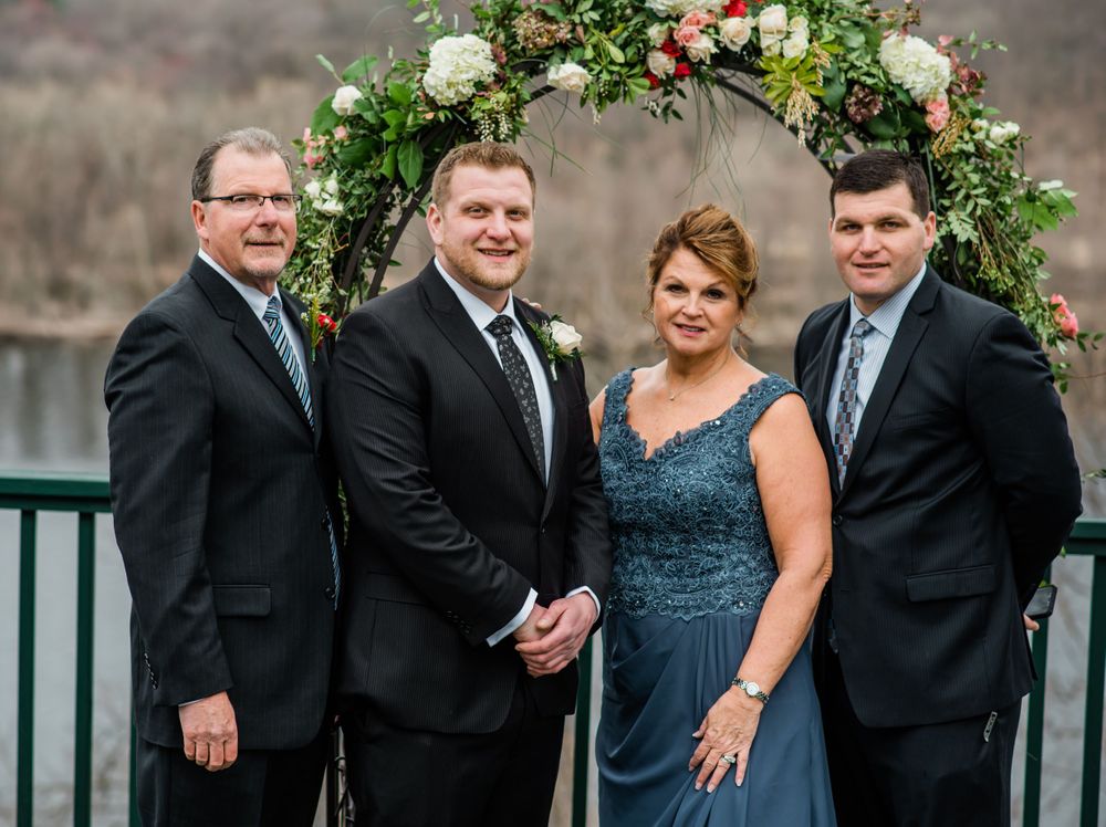 Wedding party poses under a floral arch against a mountain backdrop.