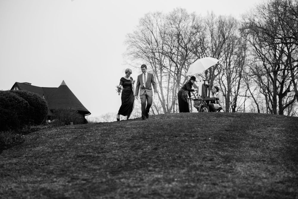 Black and white photo of wedding guests walking on a hilltop with umbrellas.