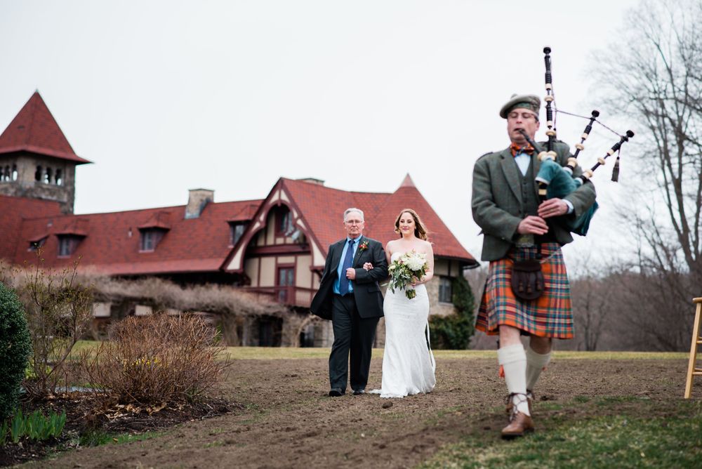 Traditional bagpiper leads a wedding processional outside a Tudor-style mansion.