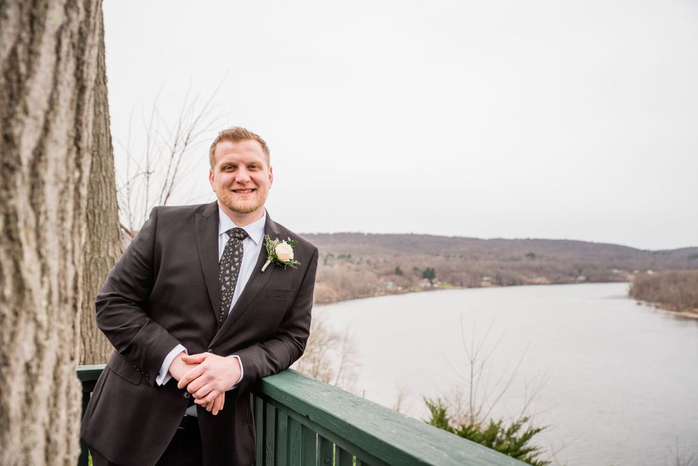 A person in a dark suit smiles while standing on a deck overlooking a lake with winter hills in the background.