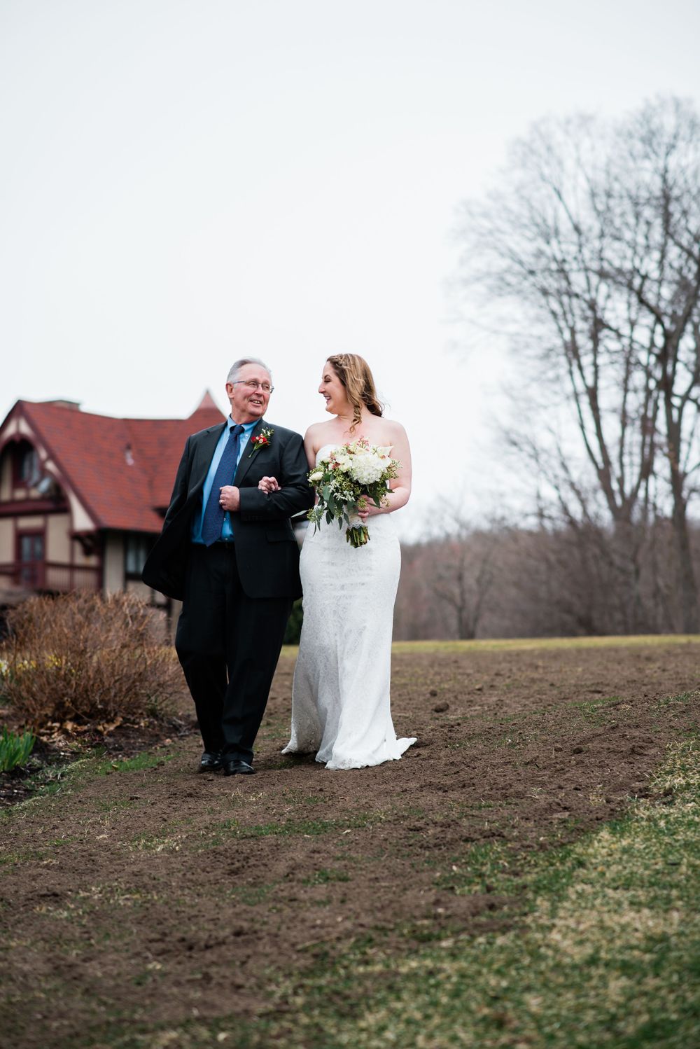 A bride in white gown walks with an escort on a hilltop path with a red house in the background.