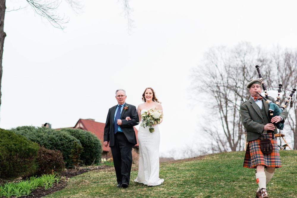 A wedding processional walks along a path with a bagpiper in traditional Scottish attire leading the way.