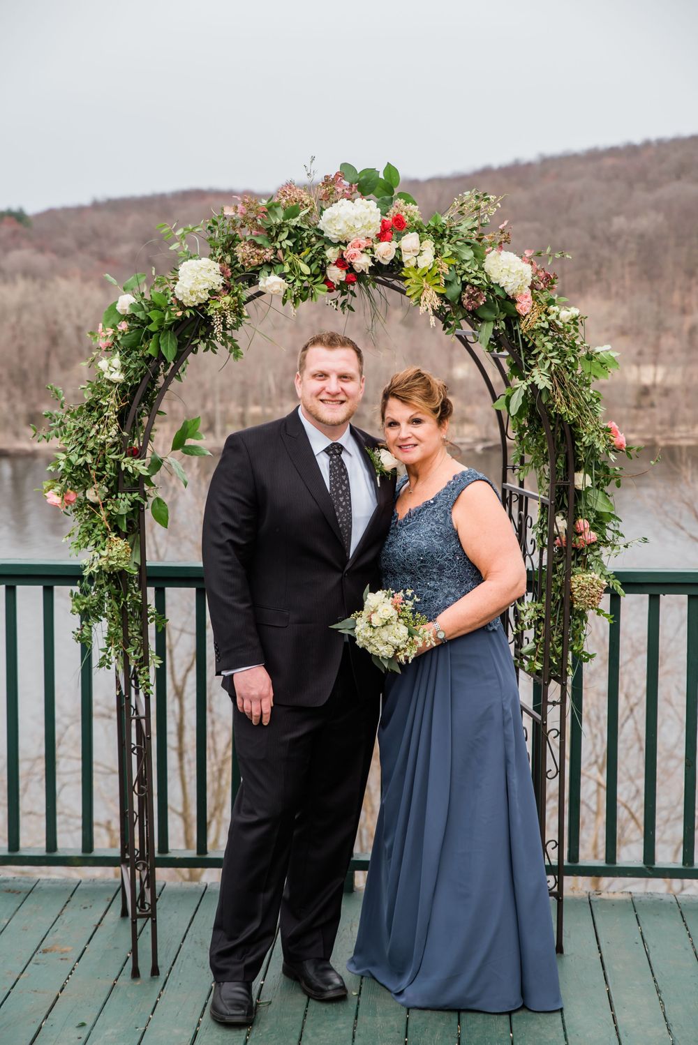 Couple stands under an elegant floral arch on a deck overlooking a scenic lake view.
