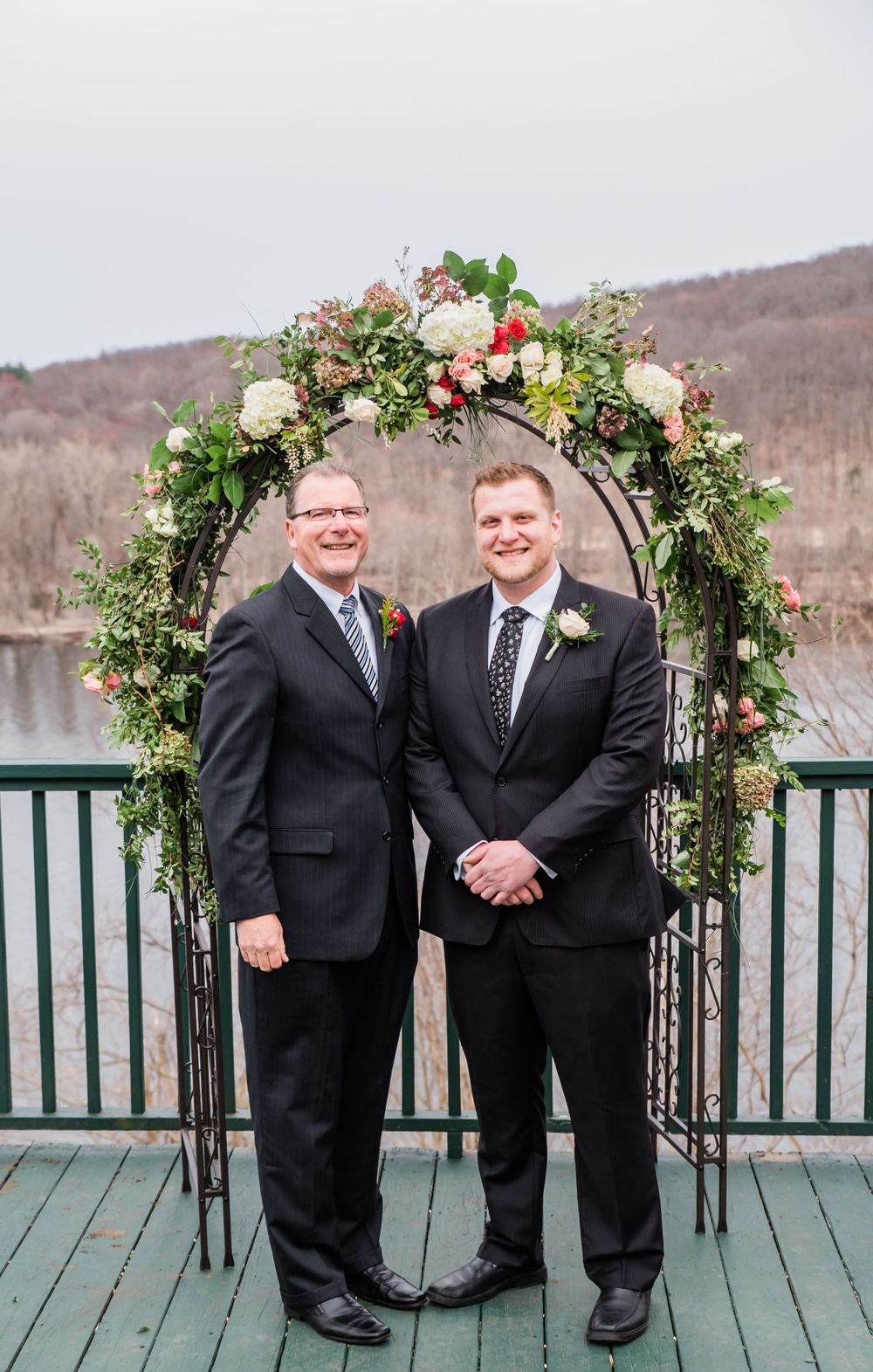 Two people pose under a lush floral wedding arch on a deck with a lake backdrop.